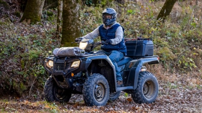 Driver on green Foreman going left on dirt road. Trees and bushes in background.