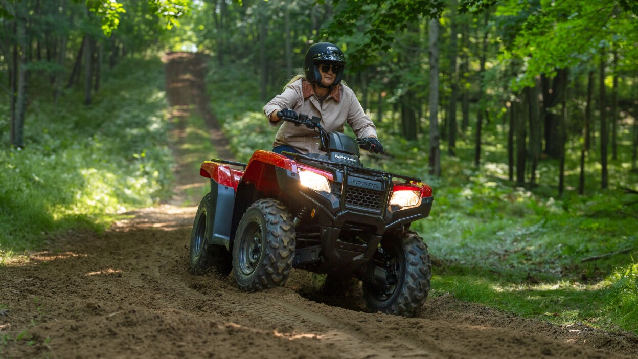 Front angle image of driver on Rancher with headlight on, on dirt road. Wood landscape in background.