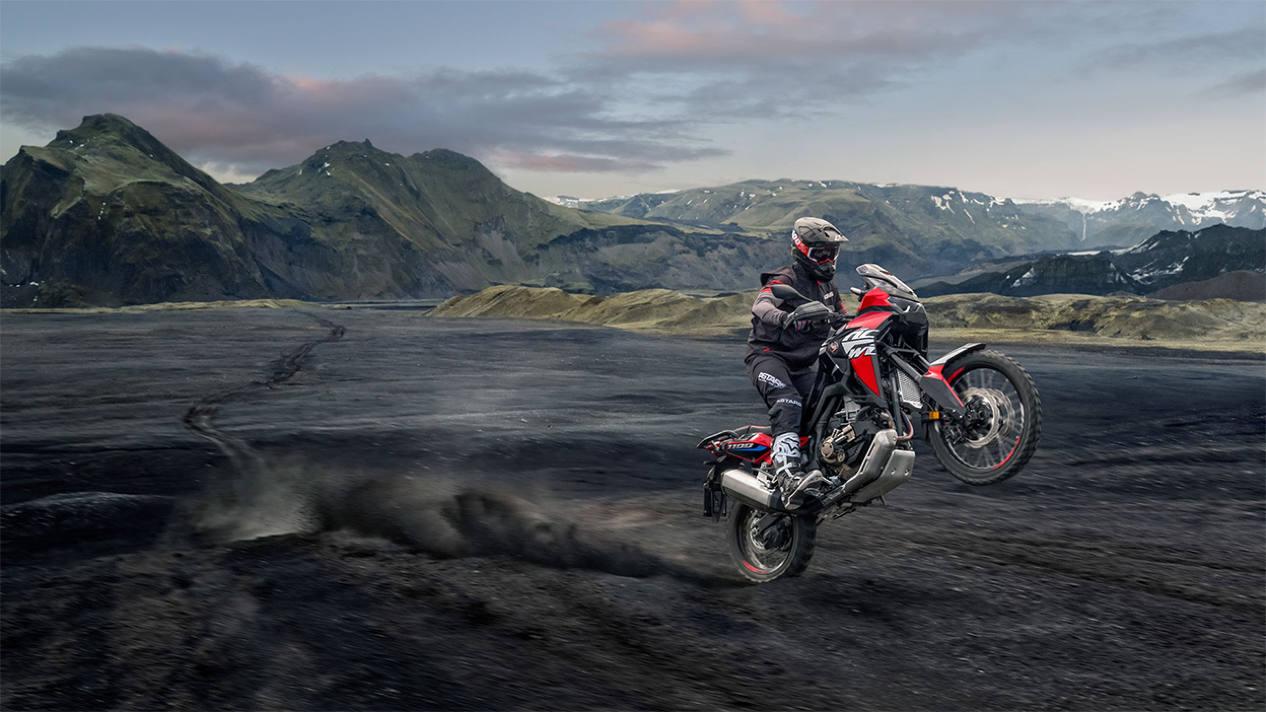 A professional rider on an Africa Twin doing a wheelie on black sand in the mountains