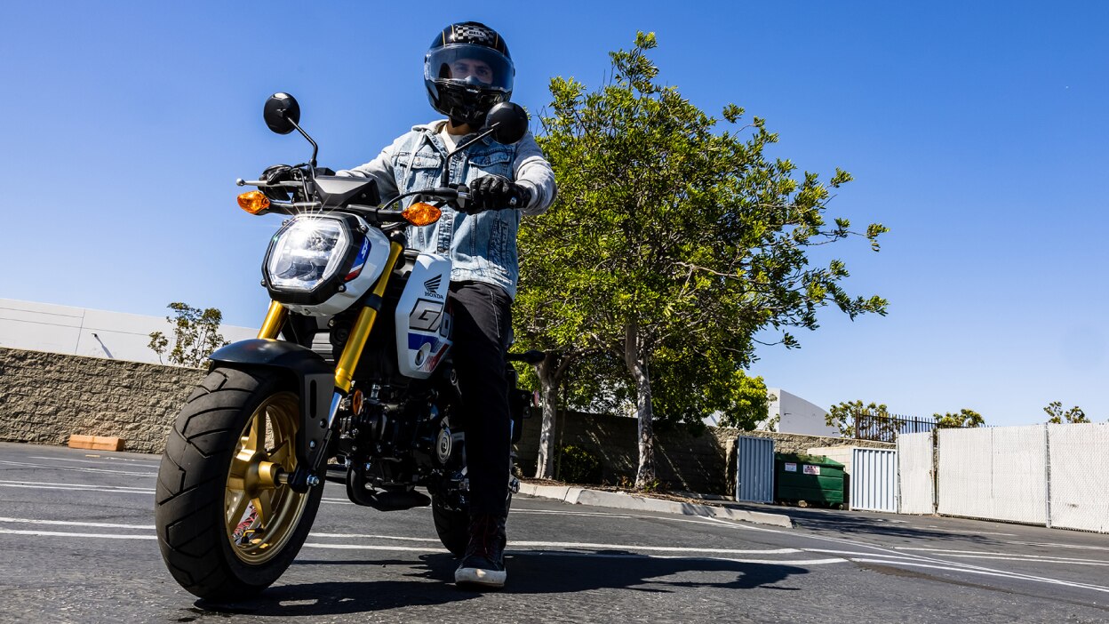 Cavalier avec veste en jean bleu clair garé sur un vélo dans un parking. Arbres, murs et ciel clair en arrière-plan.