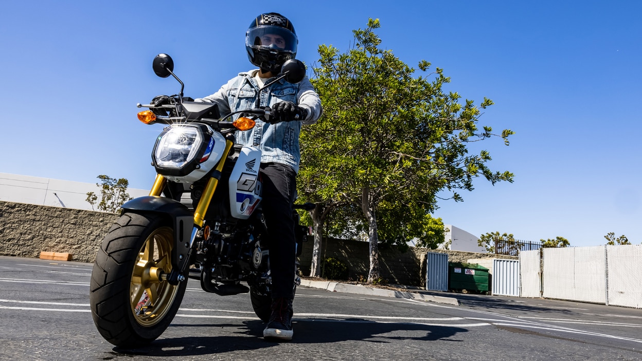 Rider with light blue denim jacket parked on bike in car park. Trees, walls and clear sky in background.