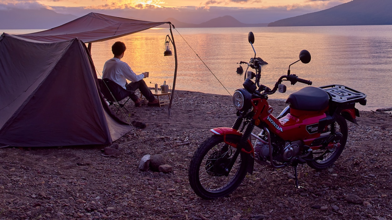 Red bike stationary at left angle. Man under tent on the left looking out at lake.