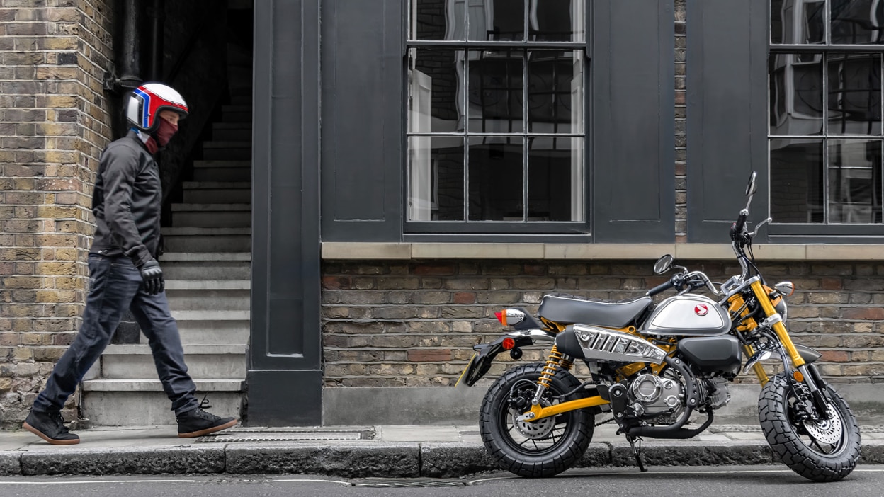 Rider with helmet on walking right towards parked yellow bike. Building with large windows in background.