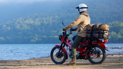 Rider with beige jacket stationary on red bike looking out onto water. Forest landscape in far background.