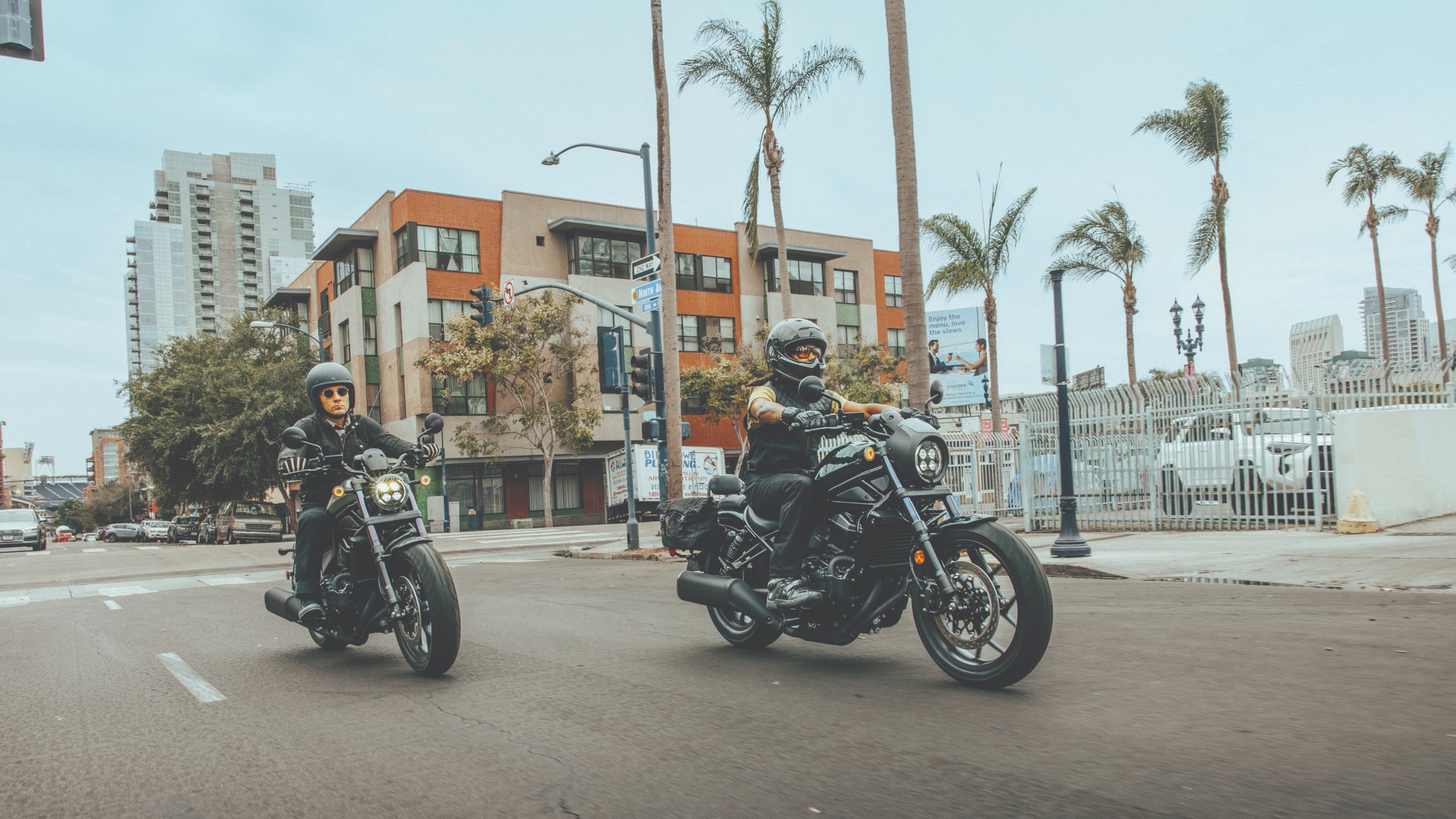 Two riders on Honda Cruiser motorcycles in an urban setting with palm trees in the background
