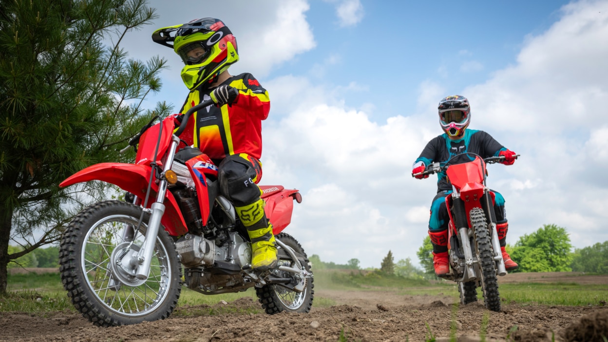 Young rider in front of adult rider on dirt track. Clear sky in background.