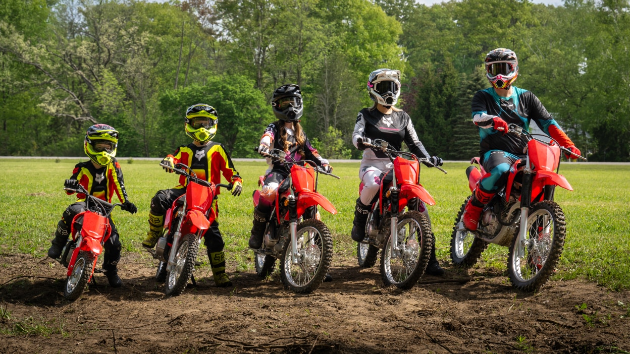 Deux cavaliers adultes, trois jeunes cavaliers à l'arrêt côte à côte. Verdure en arrière-plan.