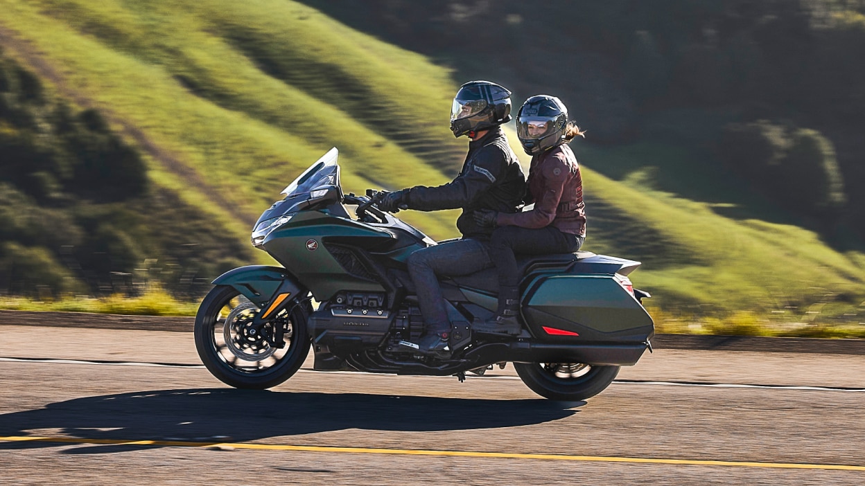 Two riders on green bike going left. Passenger looking left. Green hill in background.