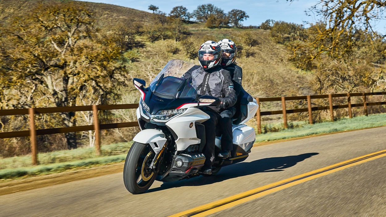 Two riders on white bike going left. Trees, hill and fence in background.