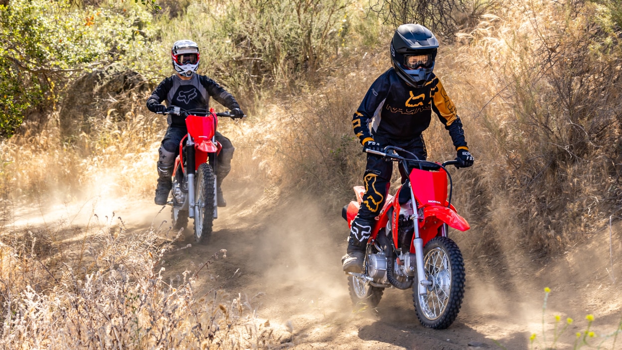 One young rider sitting up on bike on dirt track. One adult rider behind.