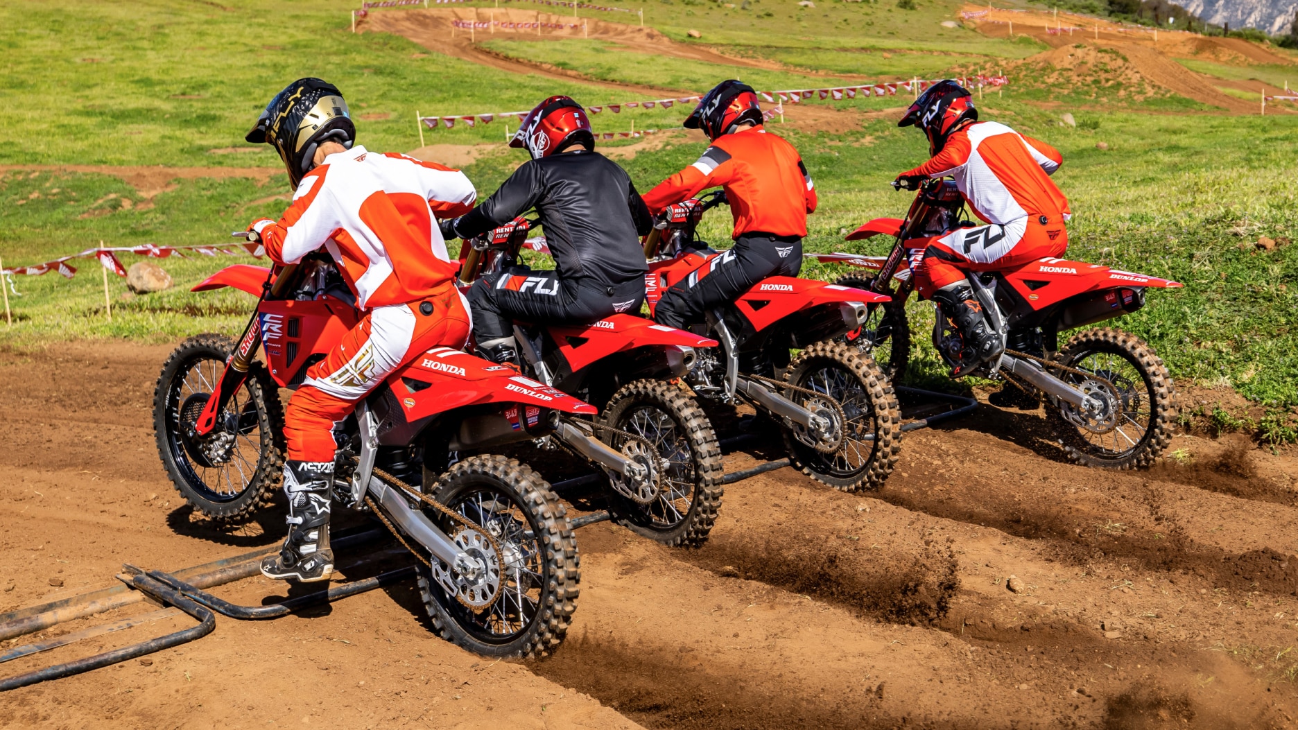 Four riders lines up about to start on dirt track. Back angle.