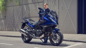 NT1100 is stationary on the road. Grey building in the background. A woman behind bike.