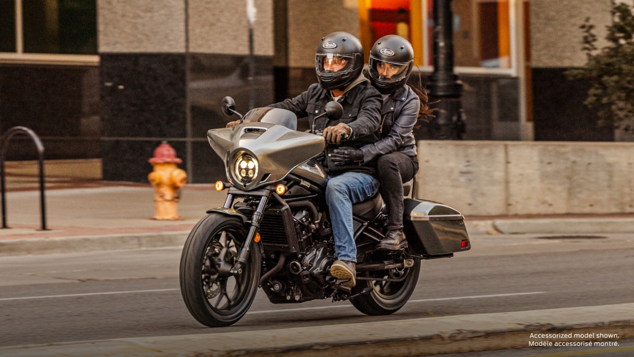 Pilote et passager sur route dans le paysage urbain. Bâtiment gris en arrière-plan. Modèle accessorisé montré.