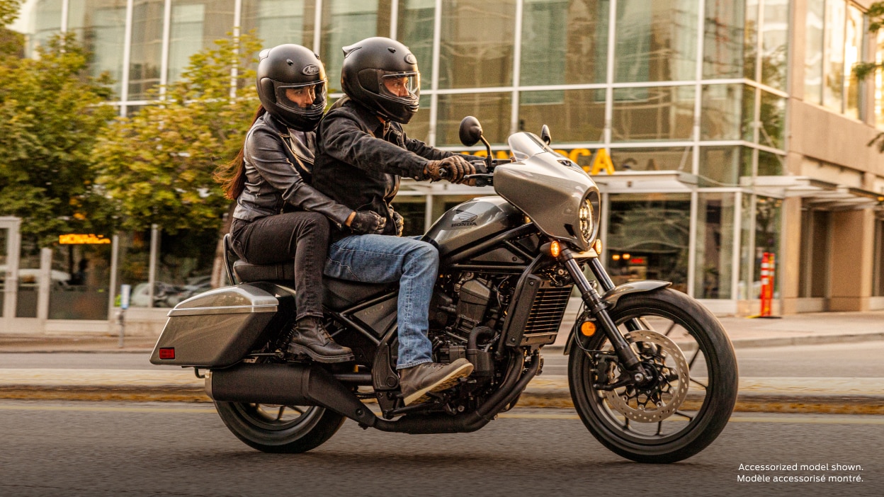 Rider and passenger going right on road. Building with large windows, side walk and tree in background. Accessorized model shown.