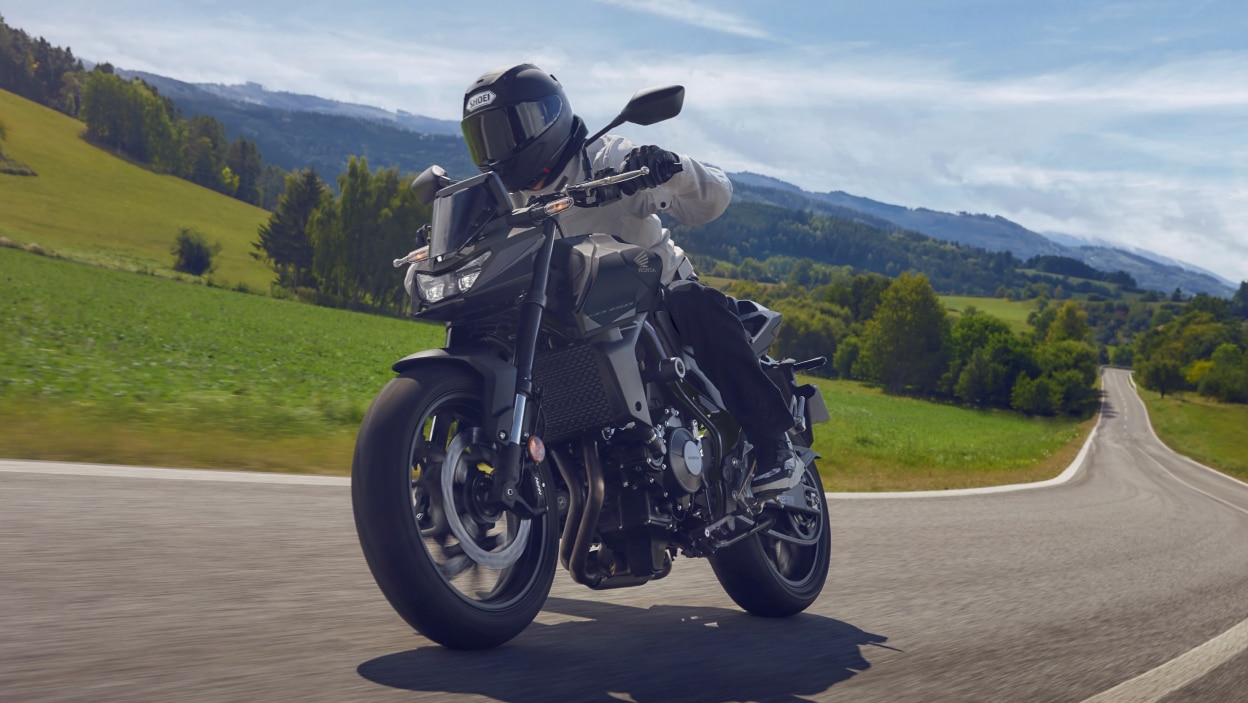Pilote sur CB750, tournant à droite sur la route, face à l'avant. Collines verdoyantes, arbres et herbe en arrière-plan