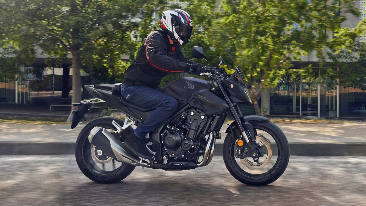 Rider on CB500F at right angle on road. Street and green tree in background.