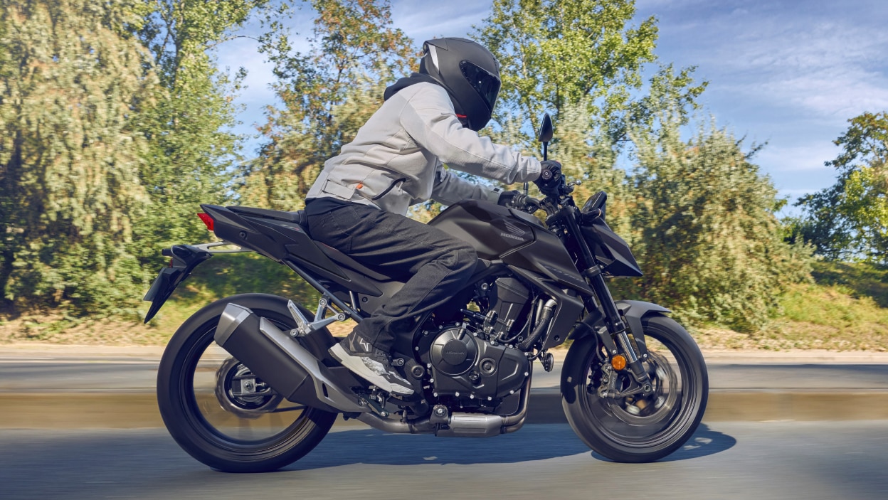 Pilote sur CB500F qui va à droite. Arbres verts et buissons en arrière-plan.