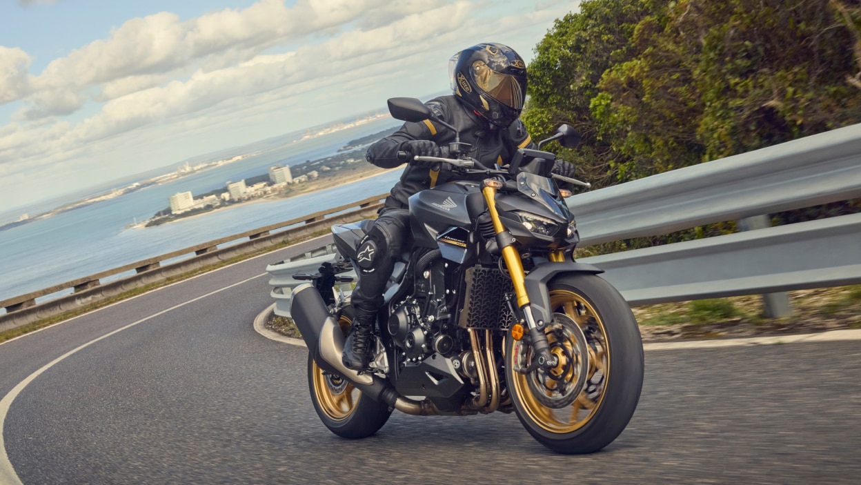 Pilote effectuant un virage à gauche sur CB1000SP. Mer et ville en arrière-plan.