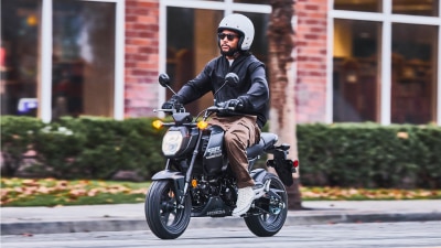 Driver with sunglasses and white helmet riding Grom through street. Building with large windows in background.