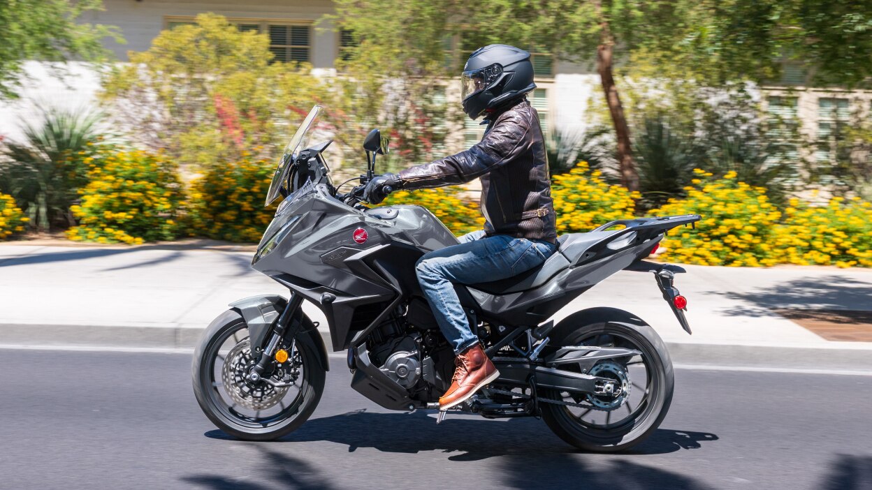 Rider on NT1100 going left on road. Pavement,  colourful bushes and small trees in background.