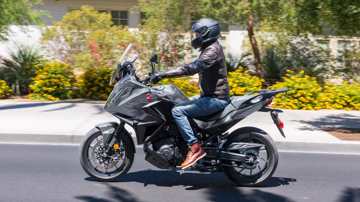 Rider on NT1100 going left on road. Pavement, colourful bushes and small trees in background.