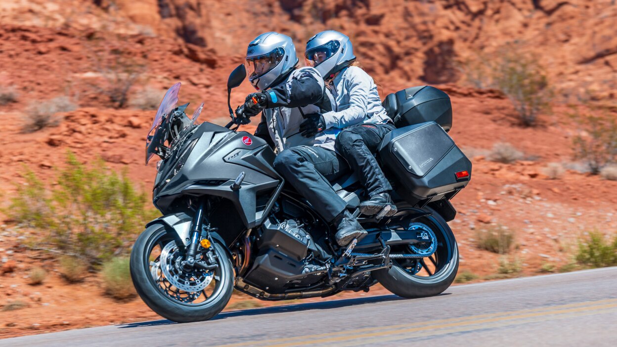 Rider and passenger on NT1100, going left on road. Red rocky landscape in background.