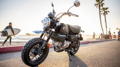 Monkey motorbike. Stationary on road. Beach and man with surf board in background.