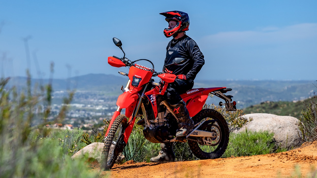 Rider stationary on CRF450L. City landscape and blue sky in background.