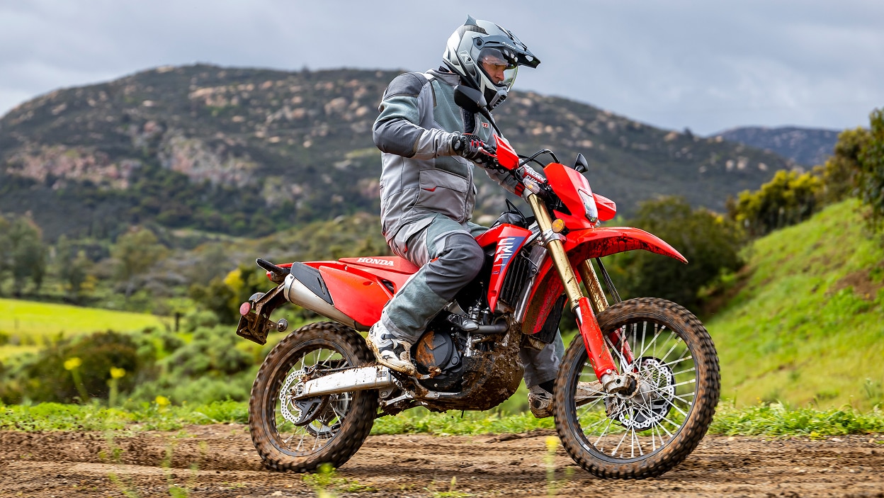Pilote sur une CRF450L sur un chemin de terre. Buissons verts en arrière-plan.