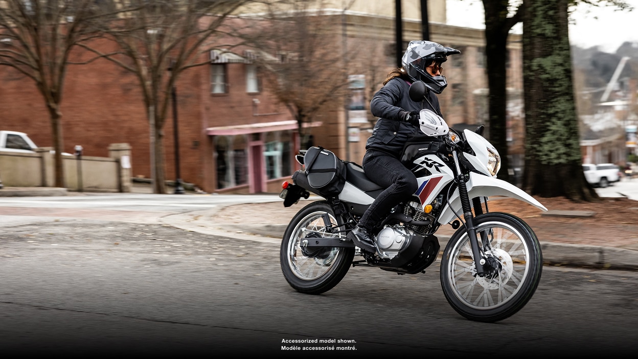 Pilote à l'arrêt sur une CRF450L. Paysage urbain et ciel bleu en arrière-plan.