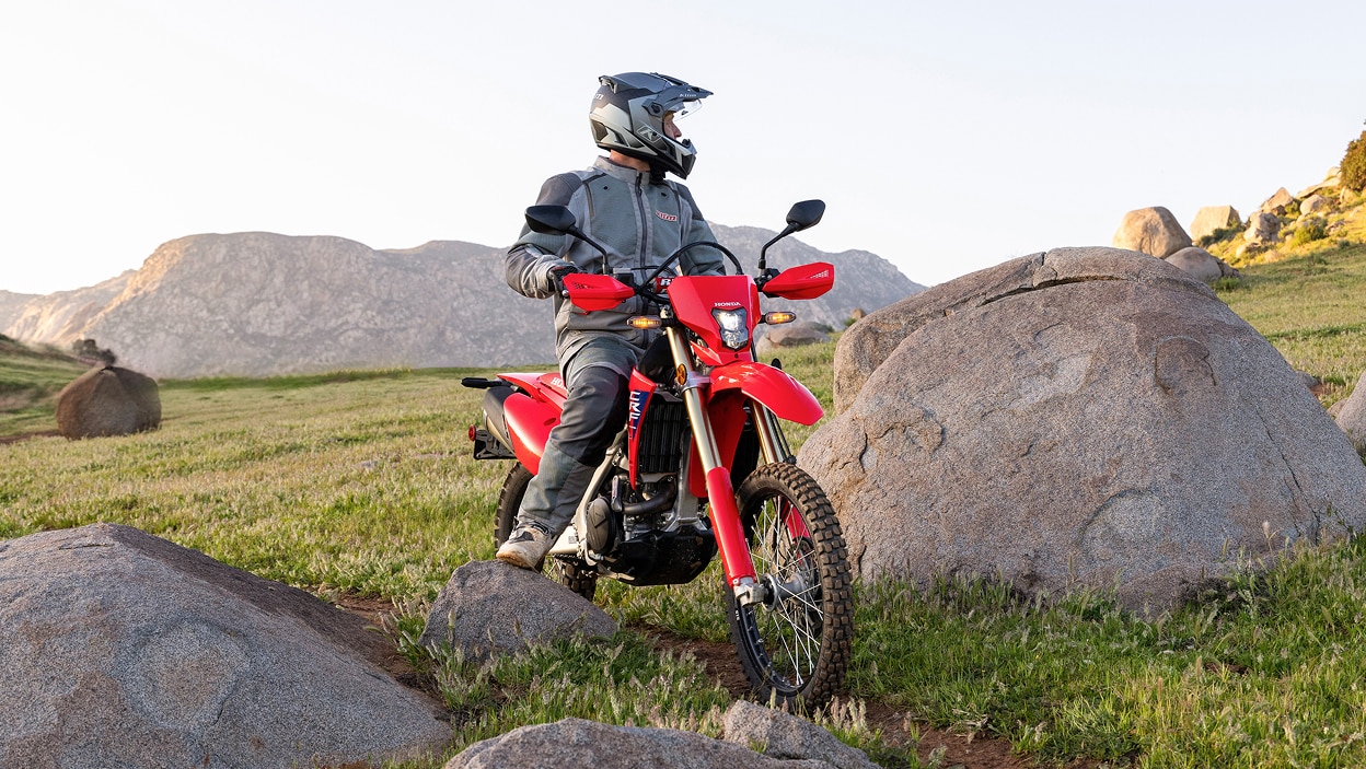 Rider standing on CRF450L. Rocky and green hills in background.