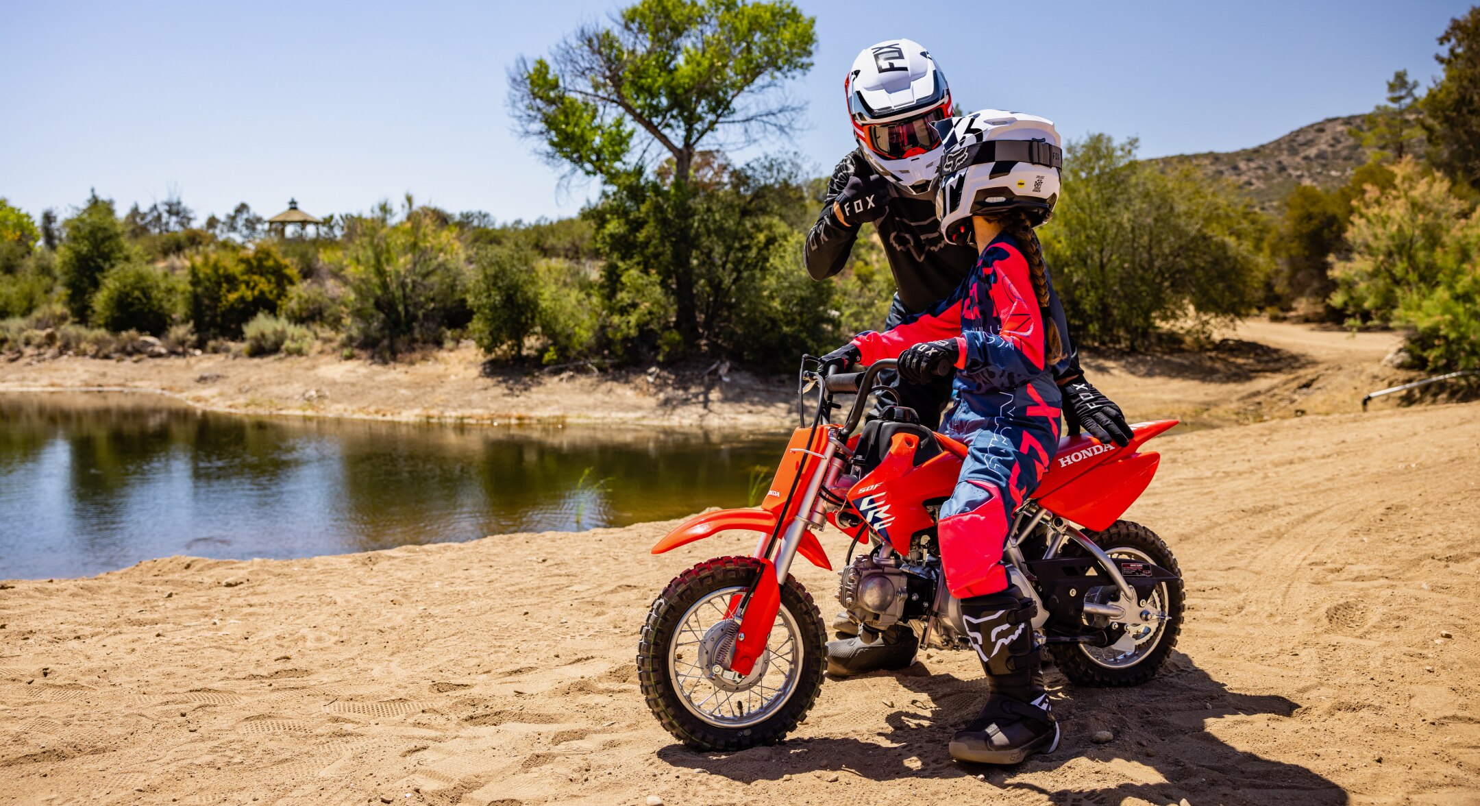 Young rider on bike with instructor on dirt track. Lake in background.