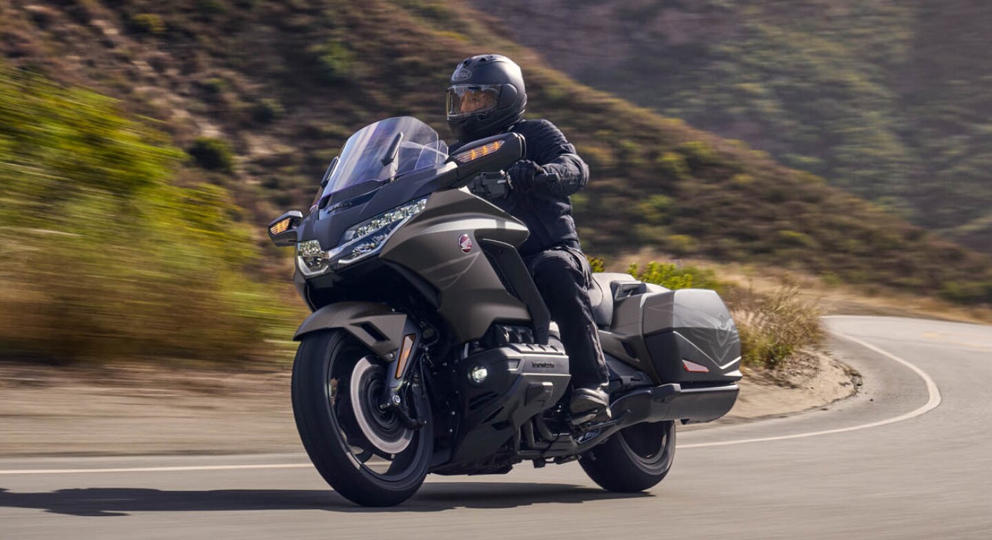 Un motard en Gold Wing tourne à gauche sur la route. Collines verdoyantes en arrière-plan.
