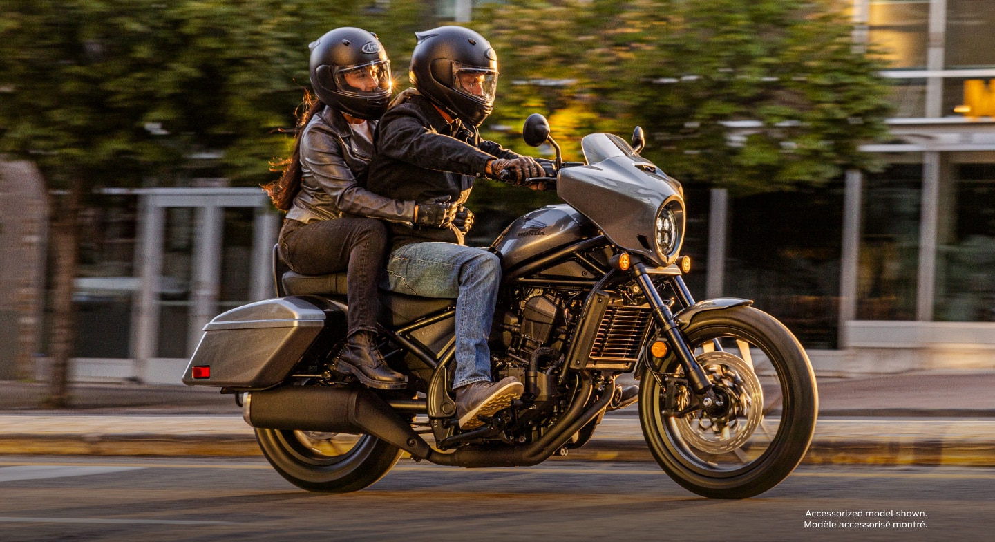 Two riders going right on street. Trees and buildings in background. Accessorized model shown.