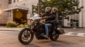 Two riders going left on street. Tree and building in background. Accessorized model shown.