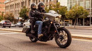 Two riders going right on street. Trees and buildings in background. Accessorized model shown.