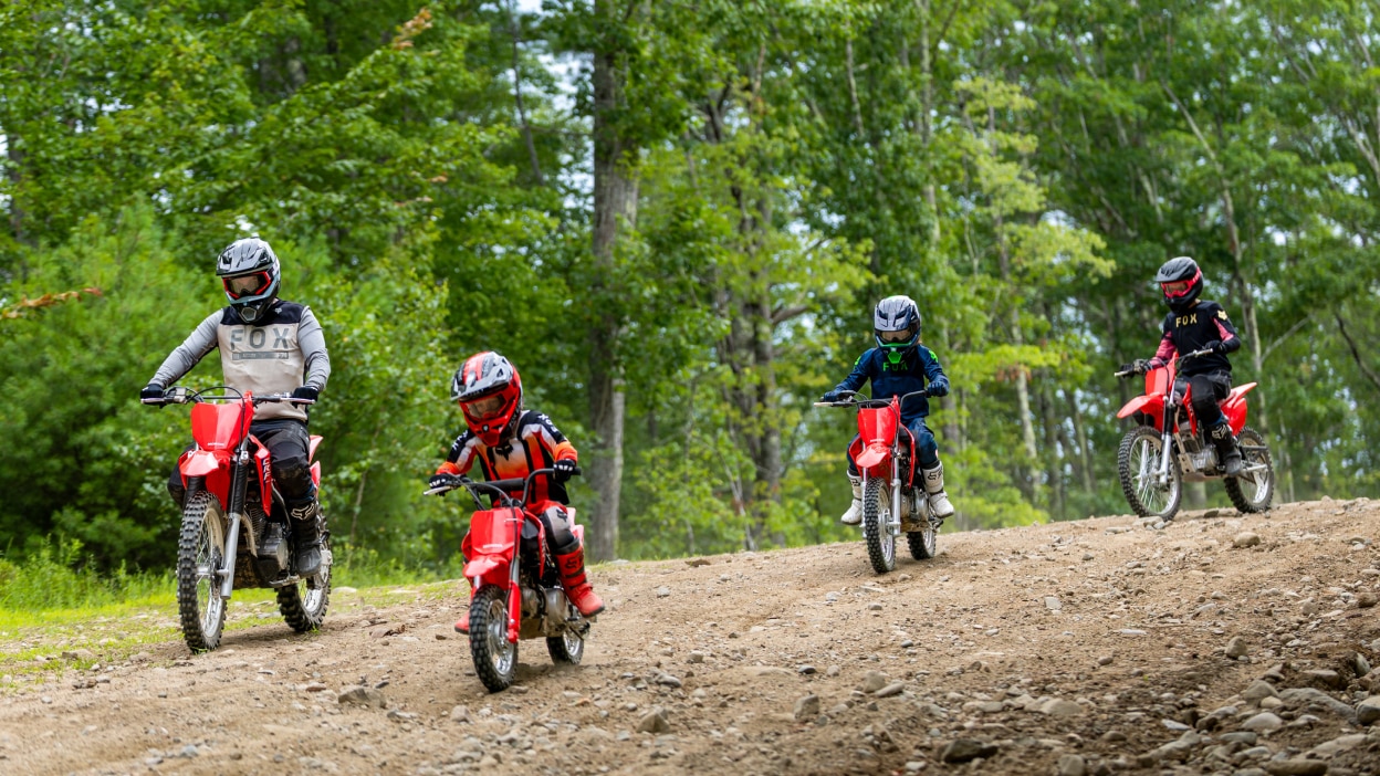Four riders on red Honda trail bikes riding toward the camera on a sandy dirt track surrounded by dense green trees.