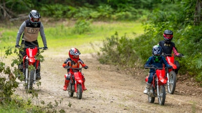 Deux jeunes conducteurs sur de petites motos tout-terrain rouges naviguant sur un sentier de terre à travers les bois.