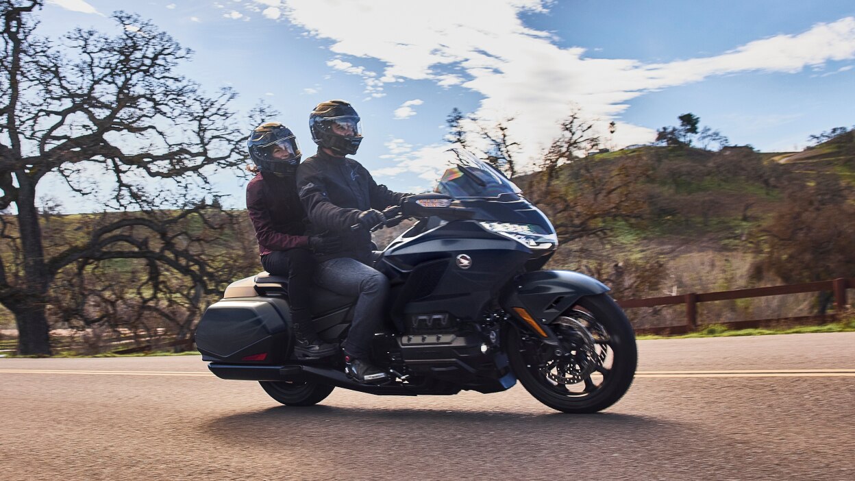 Deux personnes conduisant une moto de tourisme noire sur une route près d'arbres dénudés et de collines.