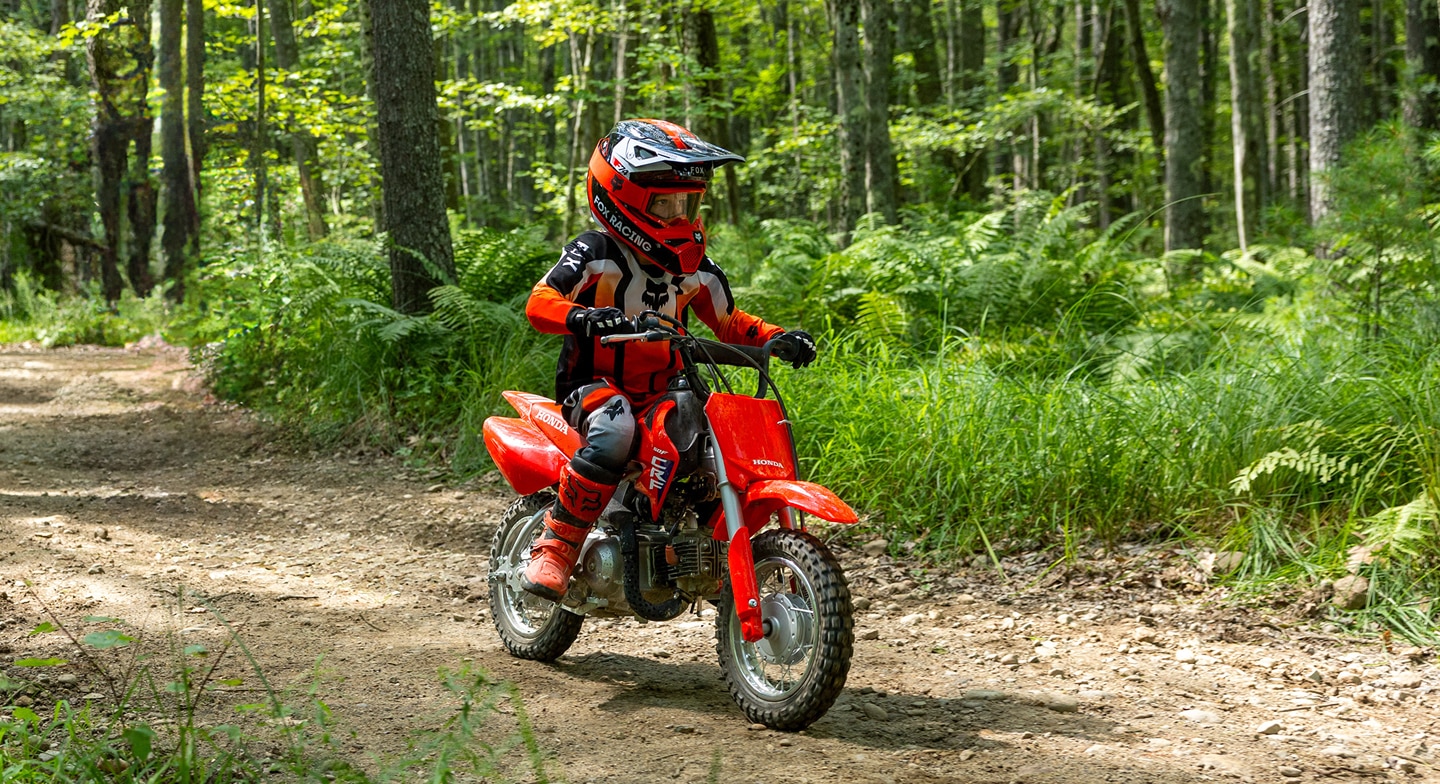 A young rider in full safety gear riding a CRF50F on a sunlit forest path.