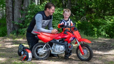 An adult and child standing next to the CRF50F, showing the durable plastic bodywork.