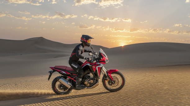 Une personne conduisant une moto sur des dunes de sable au coucher du soleil. / A person riding a motorcycle over sand dunes at sunset. 
