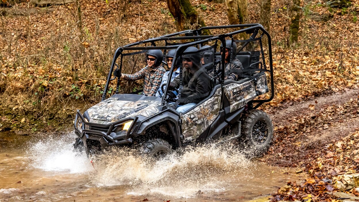 Four people in camo sxs going down in to muddy water. Leafy forrest background.