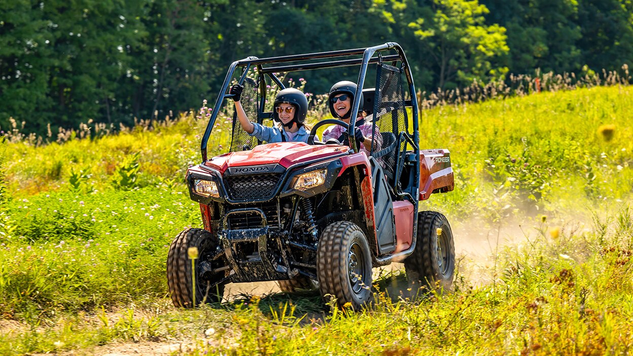 Two people driving a Honda Pioneer 520 on a grassy trail next to a field.