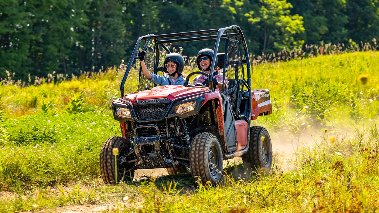 Two people driving a Honda Pioneer 520 on a grassy trail next to a field.