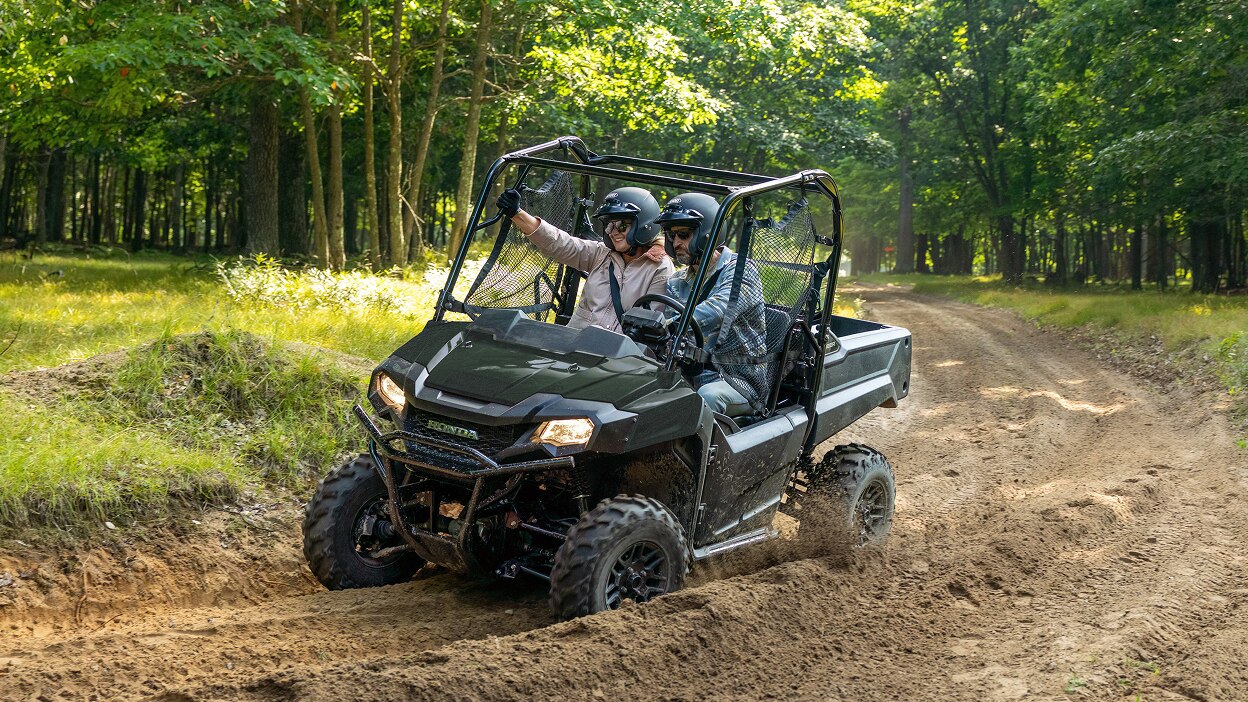 Two people driving a green Honda Pioneer 700 through a muddy forest trail.